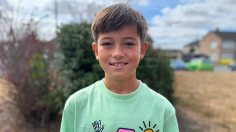 A young boy with brown hair who is smiling at the camera. He is wearing a mint green t-shirt which has a sun and a skeleton on it, and the skeleton is wearing a pink hat