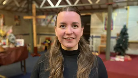 A lady with long brunet hair, she is wearing a black high necked top.