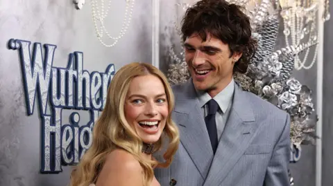 Two smiling celebrities, captured mid-laugh, stood in front of premiere branded background for the Wuthering Heights movie. To the left is blonde haired Margot Robbie looking over her shoulder. To her right is brunette Jacob Elordi looking at the back of her head.