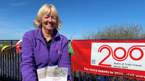 Jayne Sumner, who has blonde hair and is wearing a purple zip jumper, smiling and holding the booklets. She is on a train platform on a sunny day while standing next to a red Railway 200 banner.
