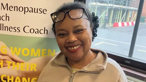 Heather is wearing her glasses on the top of her head, her hair is tied back to a bun and she is smiling at the camera, she is wearing a beige zipped jumper with a yellow top peaking out on the top. 