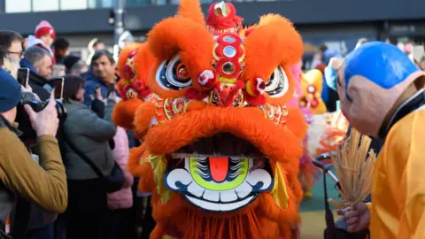 Birmingham Hippodrome A large orange lion costume fills the centre of the frame during a Chinese New Year lion dance parade. Crowds line the street on either side, many taking photos as the procession passes.