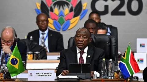 Getty Images South African President Cyril Ramaphosa seated (right) in black suit and white shirt and microphone and a tablet. In front of him on the table is a South African flag poster written "South Africa G20 President'