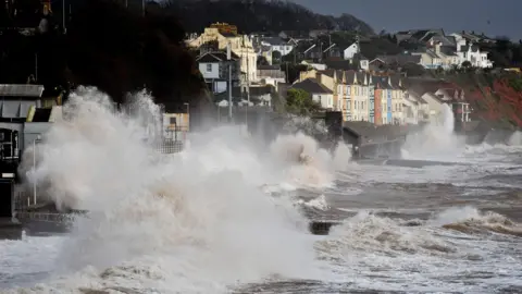 Waves break across the length of the sea wall in Dawlish. The large waves are crashing over the wall. The sea is very chopping. Houses overlook the water.