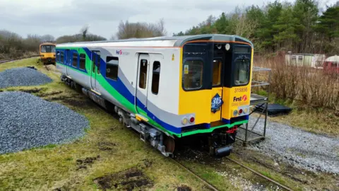 ACMU Society A picture from slightly above the Class 315 train that sits on railway tracks. Another electric train can be seen behind it in the distance along with some small outbuildings. 