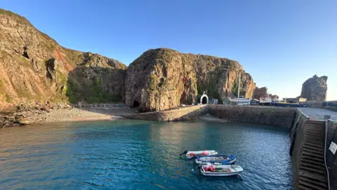 Sark Tourism Creux Harbour in Sark on a sunny day. There are three small boats on the water next to the harbour wall. A yacht is in the harbour but has been taken up onto the slipway. There is the entrance to a tunnel that cuts through cliffs next to the yacht. There is also a small beach.