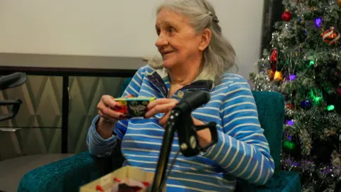 Corgi An elderly woman wearing a striped blue and white top holding a model car toy inside a box. She is sitting next to a Christmas tree.
