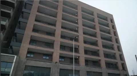 A multi-storey brick residential building with rows of balconies on each floor, viewed from street level.