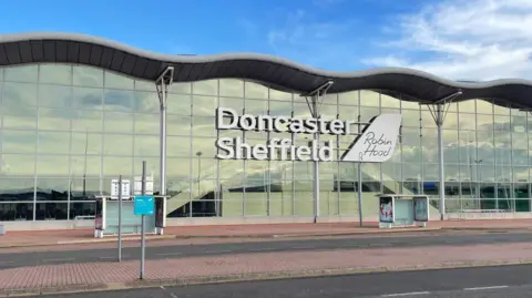 The front of Doncaster Sheffield Airport.  A glass fronted building with a wavy roof.  There is a large sign on the front of the building saying Doncaster Sheffield, with Robin Hood in smaller lettering to the side.