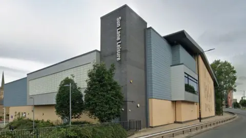 A grey and sand-coloured building with the words Sun Lane Leisure on. The building is set on the roadside with trees and green bushes around the side. 