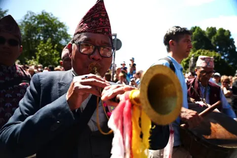 Close-up of a parade musician playing a long brass horn decorated with colourful ribbons while fellow musicians and a crowd of spectators gather along the parade route on a bright summer day.
