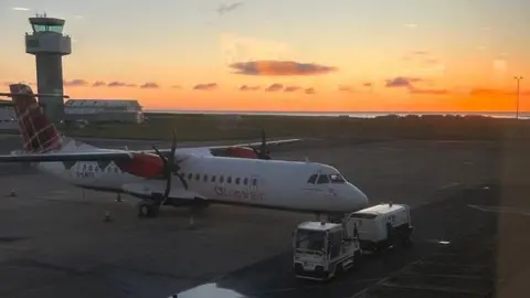 BBC A Loganair plane on the tarmac at Ronaldsway at sunrise. There is a small vehicle for transporting luggage parked in front of it .