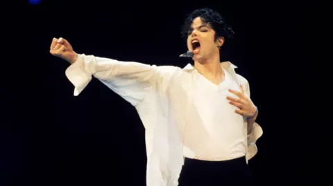 Getty Images Michael Jackson is on stage wearing a white undershirt and white shirt, with a microphoned strapped to the side of his face. He is shouting or singing. It is dark around him.