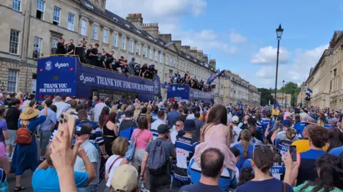 Fans in blue black and white waving flags with two blue open top buses passing in the background with players on top waving