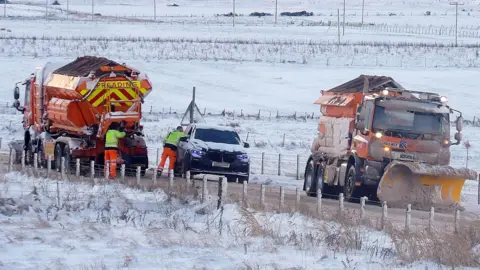 BBC Weather Watchers/Standing Stone Two snow ploughs and a blue car in the middle of them on a country road with snowy fields