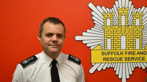 Chief fire officer Jon Lacey standing in front of a red wall with the Suffolk Fire and Rescue Service logo 