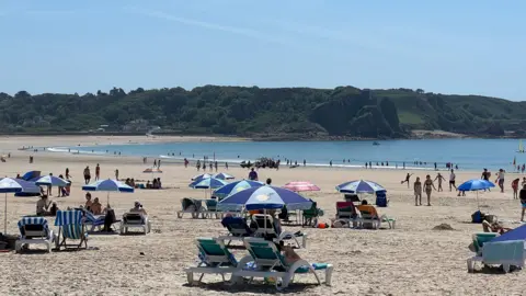 BBC A picture of a sunny St Brelade beach. It shows people relaxing under umbrellas and walking along the shore. The sea and green cliffs are visible in the background.