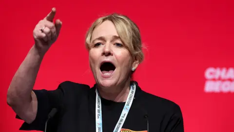 A woman with blonde hair tied back in a ponytail stands in front of a red background. She is wearing a black top and has a lanyard round her neck. She is gesticulating with her hand raised and forefinger pointing and she appears to be shouting or at least talking animatedly.