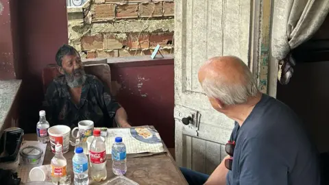 Matthew Gayle Nizam Hosein speaking to Ian McKay at Hosein's house in Trinidad. They both sit at a table filled with plastic bottles of water and coca cola. It is a porch with open windows on either side and is quite run down. Nizam wears an open necked dark shirt with paisley patterns and has a long bushy beard and short dark hair. Ian is seen from behind wearing a blue/grey t-shirt with red sunglasses clipped onto it.