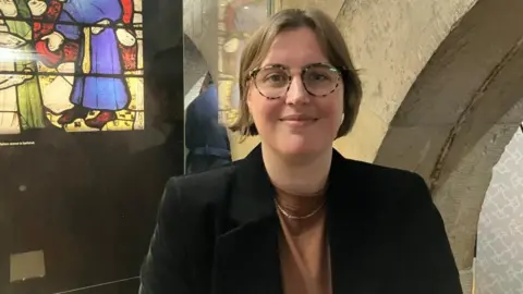 Dr Jennie England stands in front of glass display cabinet, with a depiction of colourful stained glass behind her. She has short brown hair and wears tortoiseshell glasses, a black blazer and a brown top. 