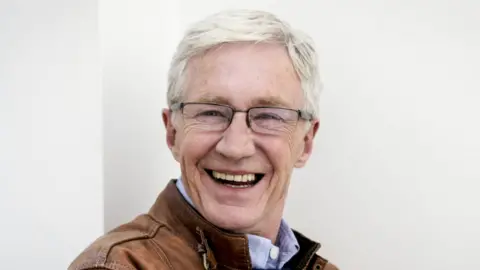 O'Grady smiles at the camera in front of a white wall. He is silver-haired, the picture taken a few years before his death.