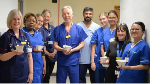 A group of doctors in blue scrubs stand looking at the camera and smiling. They are all holding small white containers.