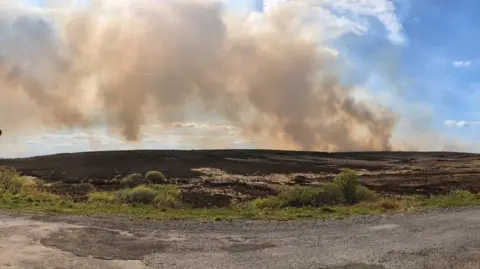 A wide shot of a large plume of smoke over a mountainous area in Clogher. There are blue skies with some white clouds. 