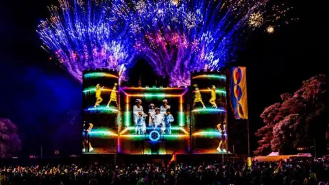Camp Bestival It's dark and a crowd of thousands surround Lulworth Castle watching fireworks and illuminations. The symmetrical keep has a round tower on each corner and videos of people are projected on to the stone walls. Purple and pink fireworks explode from the turrets.