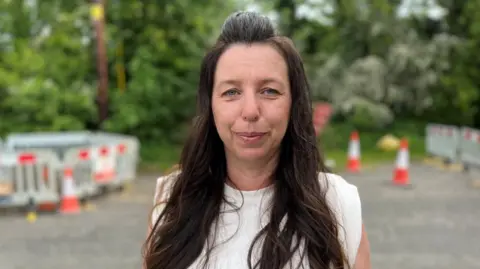 A woman with long brown hair, wearing a white sleeveless top looks at the camera. There are roadworks and traffic cones behind her