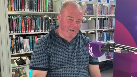A man sits in front of a purple microphone inside a library. A bookshelf is in the background. 