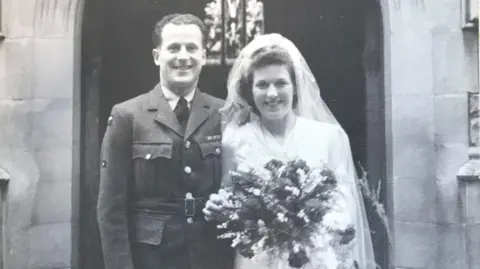 Hilda Bond A black and white photograph of Mrs Bond and her husband stood outside a church on their wedding day. Hilda is in a traditional white bridal dress holding a bouquet of flowers and her husband is wearing military uniform