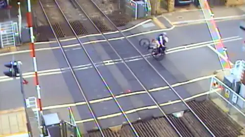 Network Rail Two cyclists are falling next to each other on a level crossing, close to the track and a lowering barrier