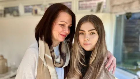 BBC/ Nicola Rees A middle aged, white woman with brown hair holds a younger woman in her arms and looks at her fondly.  They are standing in a home by a window. The younger woman has lighter brown hair and brown eyes.