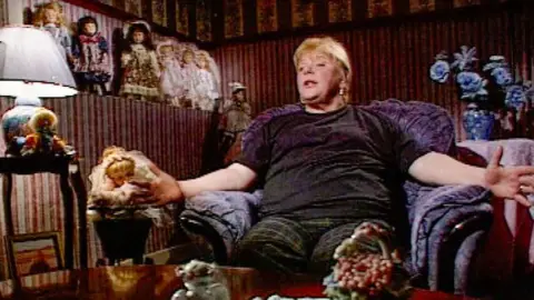 A TV image of Haney in her home from a documentary in the early 2000s. She is sitting in a big comfy purple armchair surrounded by children's dolls in old-fashioned outfits