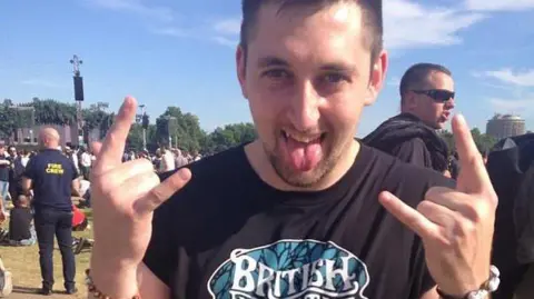 Kyle Kitchen in a Facebook image at a festival. He is wearing a black T-shirt with a logo on, has his tongue out and is making rock'n'roll horn fingers