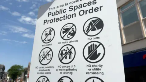 A white sign with black writing and symbols. The title of the sign is Public Spaces Protection Order. It is set against a blue sky with light clouds and there is a building behind the sign.