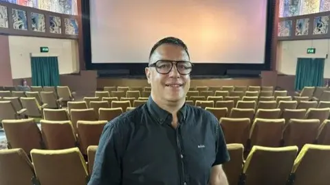 A man with dark hair, glasses and a black shirt standing in a cinema with rows of seats and a large screen behind them