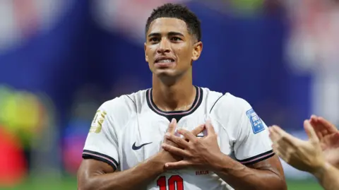 Reuters Jude Bellingham, wearing an England shirt, holds both his hands up to his chest while he is on the pitch.
