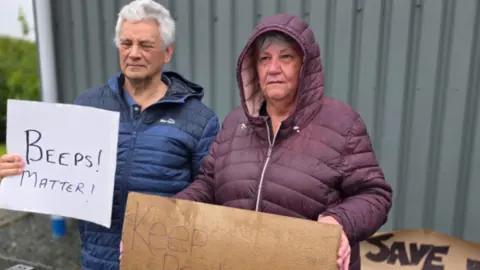 Two people stand outside an industrial building holding homemade signs reading “Beeps Matter!” and “Keep Production Local,” while another sign behind them says “Save Our Site!”.
They are standing by a roadside barrier in wet weather, suggesting a small demonstration, with the signs asking drivers to show support and highlighting concerns about the factory’s future.