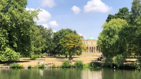 Weather Watchers/Sunshine Dan Pitville Park in Cheltenham, Gloucestershire. There is a large beige building with a green dome roof in the distance. There is a body of water and trees dotted around the park. People can be seen sat on benches in the sun and shade.
