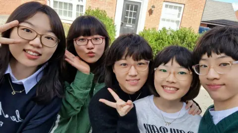 Family photo (L-R) Sisters Juliana, Clara and Ella posing for a picture with their younger siblings Ariel and Elias. Some of them are making the peace sign. They are outside in front of a house.