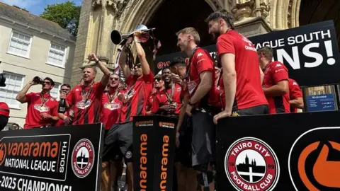 The team lift the trophy while standing on the steps of Truro Cathedral