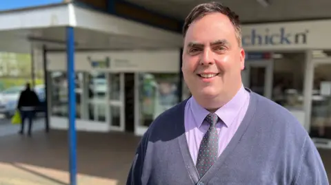 Simon Dedman/BBC Kerry Smith wearing a cardigan, purple shirt and spotted tie. His hair is dark brown. He is smiling. He is standing outside in front of a parade of shops.