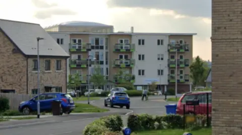 Google Clement House, a four-storey block of flats on a new-build estate. There are cars parked outside. In the foreground are houses.