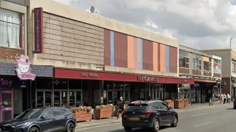 A high street with a pub exterior of The Claireville pub which had dark red painting and silver lettering. Two cars are driving past and there a number of cars parked one each side of the road.