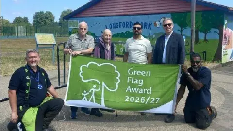A group of people stand at a park holding a large green banner. The text Green Flag Award 2026/27" is written on the front.