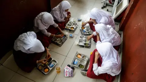 Six Indonesian girls in school uniform seated on the floor, eating their free meals
