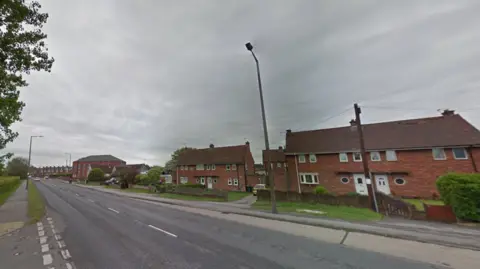 Woodhouse Green in Thurcroft. The road is quiet, with a number of semi-detached red brick homes on the far side of the road. The sky is overcast.