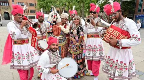 Pacemaker A group of people wearing white dresses and pink trousers. They are playing a mixture of musical instruments. 