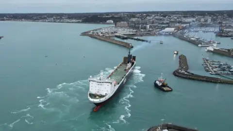 A large ferry leaving St Helier Harbour after berthing trials with a tug boat stationed nearby